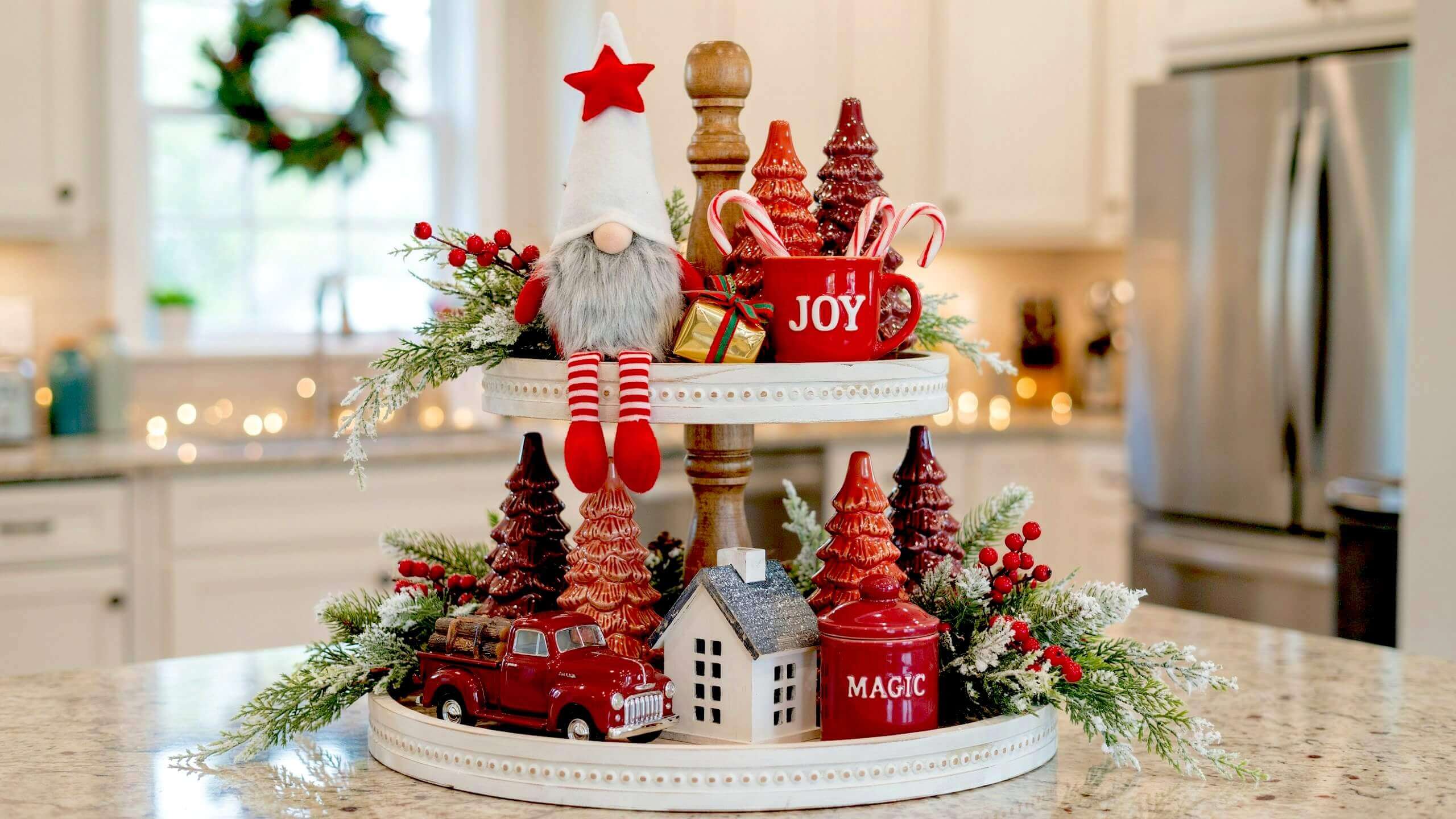 Rustic wooden two-tier stand holding a plush Christmas gnome beside red ceramic trees, a vintage toy truck, and frosted greenery for Christmas tiered tray ideas.