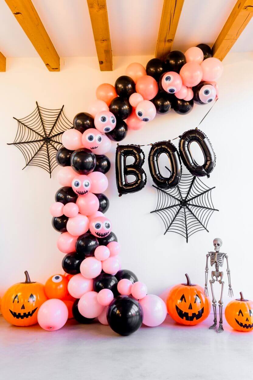 Pink and black balloons form a playful arch with spiderwebs, pumpkins, and a skeleton tucked below. This fun setup is a lively take on pink Halloween decor ideas for parties and gatherings.
