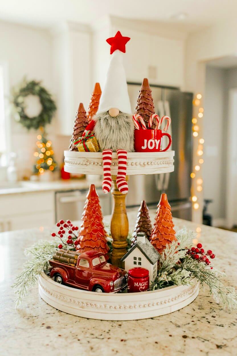 Distressed white wood stand displaying a plush grey-bearded gnome, glossy red ceramic trees, a vintage red truck model, and frosted greenery for festive tray decor.