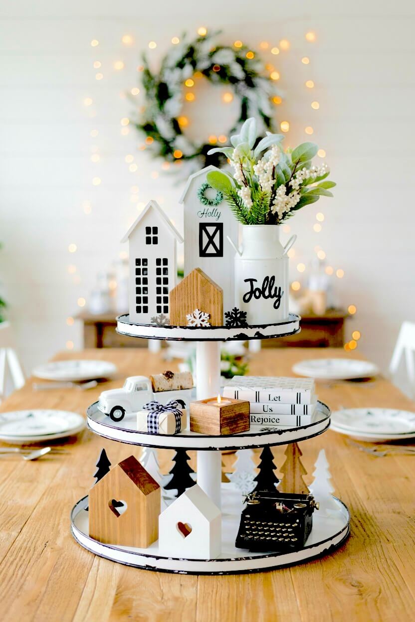 White tiered stand featuring black and white wooden farmhouse structures, a miniature vintage typewriter, and snowy artificial foliage for winter table styling.