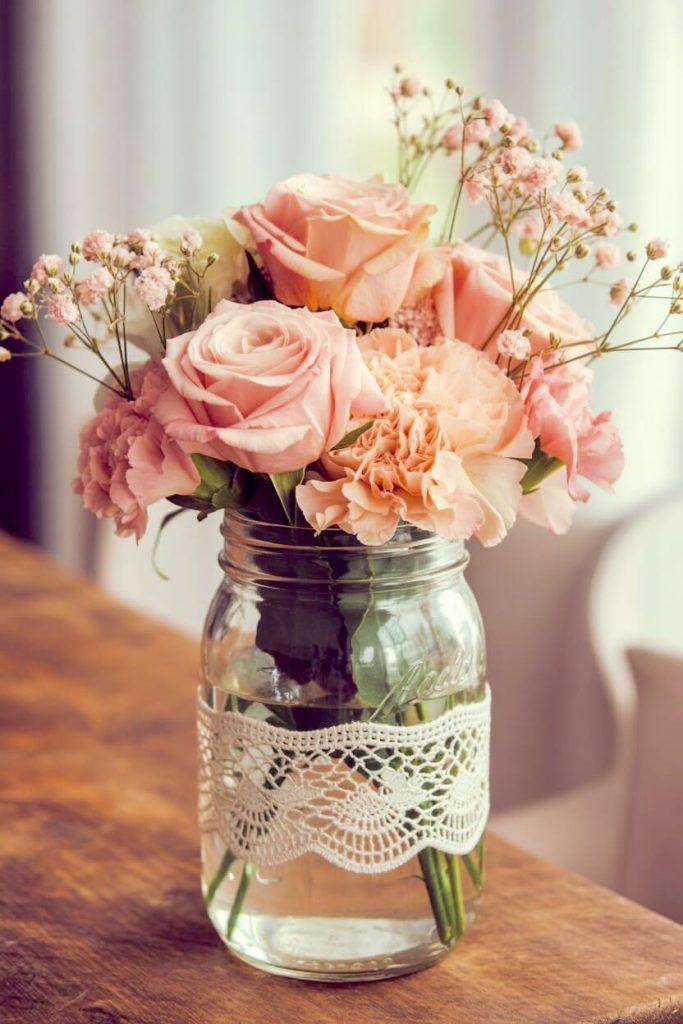 A vintage book is folded into a circular fan shape with a paper rose on top as a creative centerpiece. Three lit pink candles in glass jars and a floral teacup surround the book on a wooden tabletop. This styling focuses on recycled materials and classic floral patterns to add a literary touch to your home office or living room.