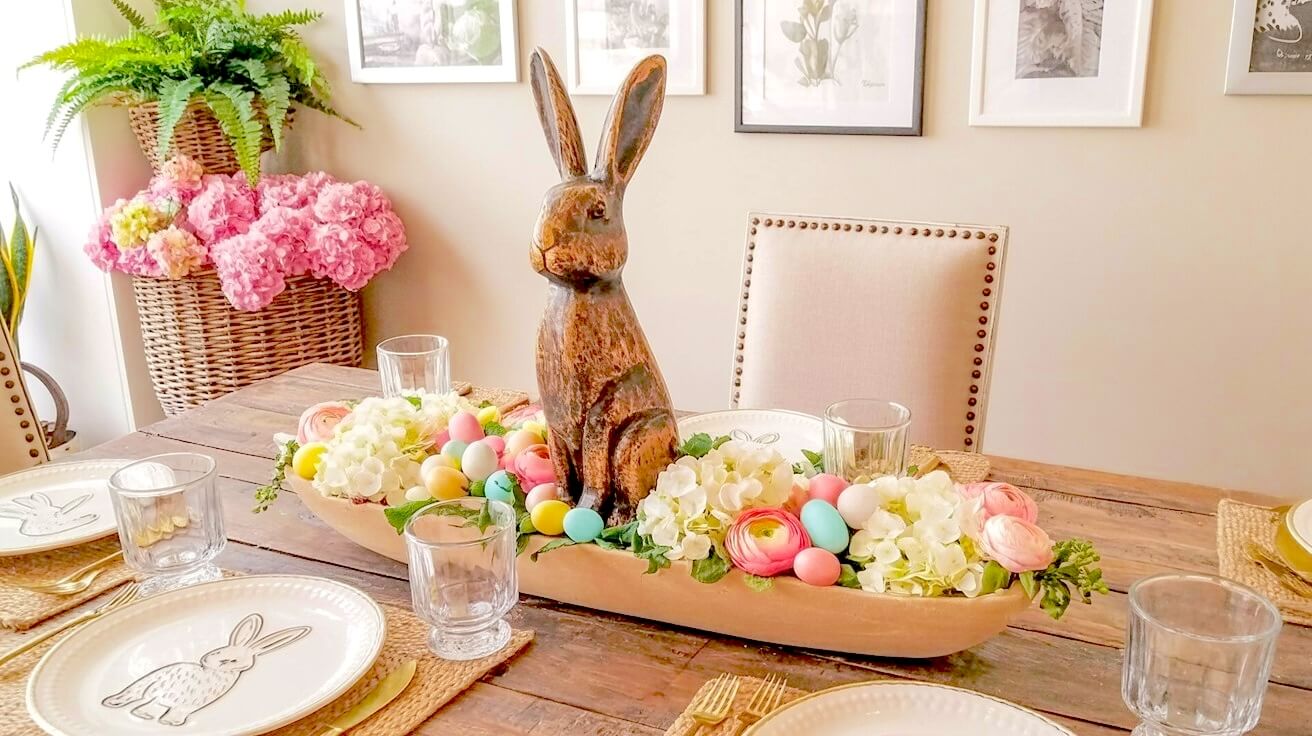An elegant Easter dough bowl centerpiece idea featuring a tall weathered wooden bunny surrounded by lush pink and white florals and colorful pastel eggs on a rustic dining table.