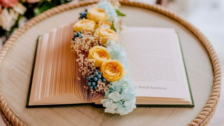 Image featuring a book centerpiece idea : a romantic top-down flat lay photograph of an open vintage book with aged pages. A beautiful arrangement of yellow ranunculus roses, pale blue hydrangeas, small blue berries, and delicate dried filler flowers is nestled along the spine. The book rests on a rustic round wooden surface with a braided rope edge. The words 'floral harmony' are visible on the right-hand page.