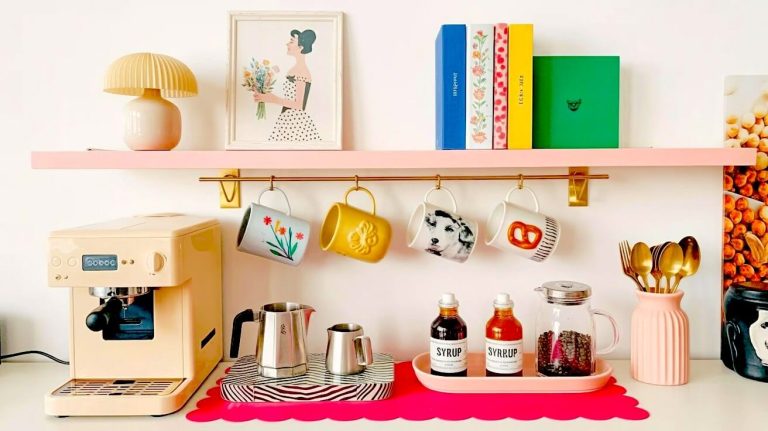 A peach espresso machine sits on a bright pink scalloped mat beneath a pastel shelf displaying bright books and art for eclectic home coffee station ideas.