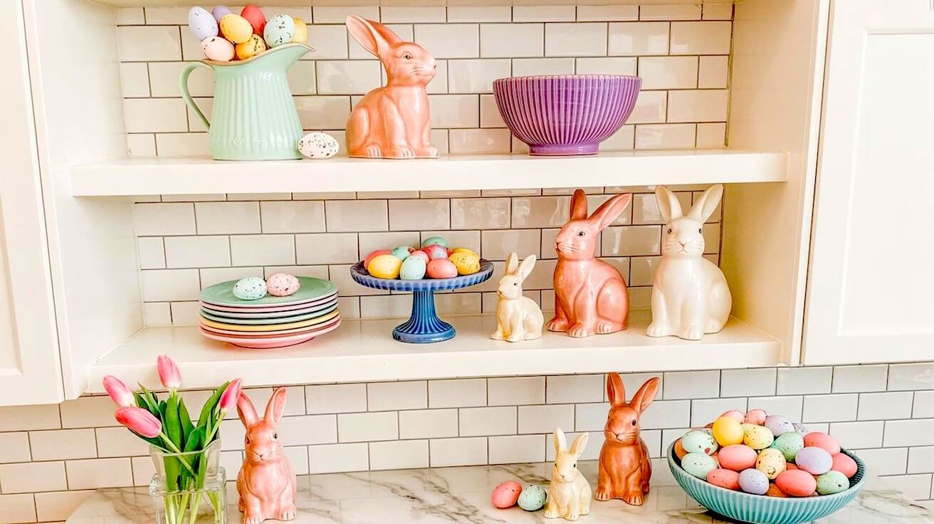 Wide shot of white kitchen shelving decorated with pastel ceramic bunnies speckled eggs and pink tulips for easter kitchen decor ideas.