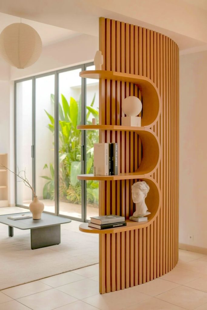 An architectural curved light wood slat room divider features built in rounded shelving displaying white ceramic sculptures and stacked books.