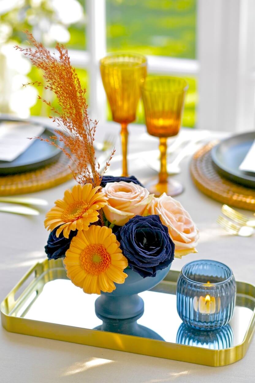 A gold mirrored tray displays a blue pedestal bowl filled with yellow gerbera daisies and deep indigo roses next to a glass candle for gorgeous autumn styling.