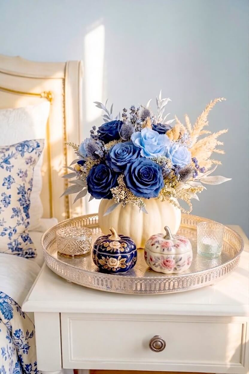 A silver tray on a nightstand holds a pumpkin vase filled with blue roses beside delicate ceramic chinoiserie pumpkins for chic autumn room styling.