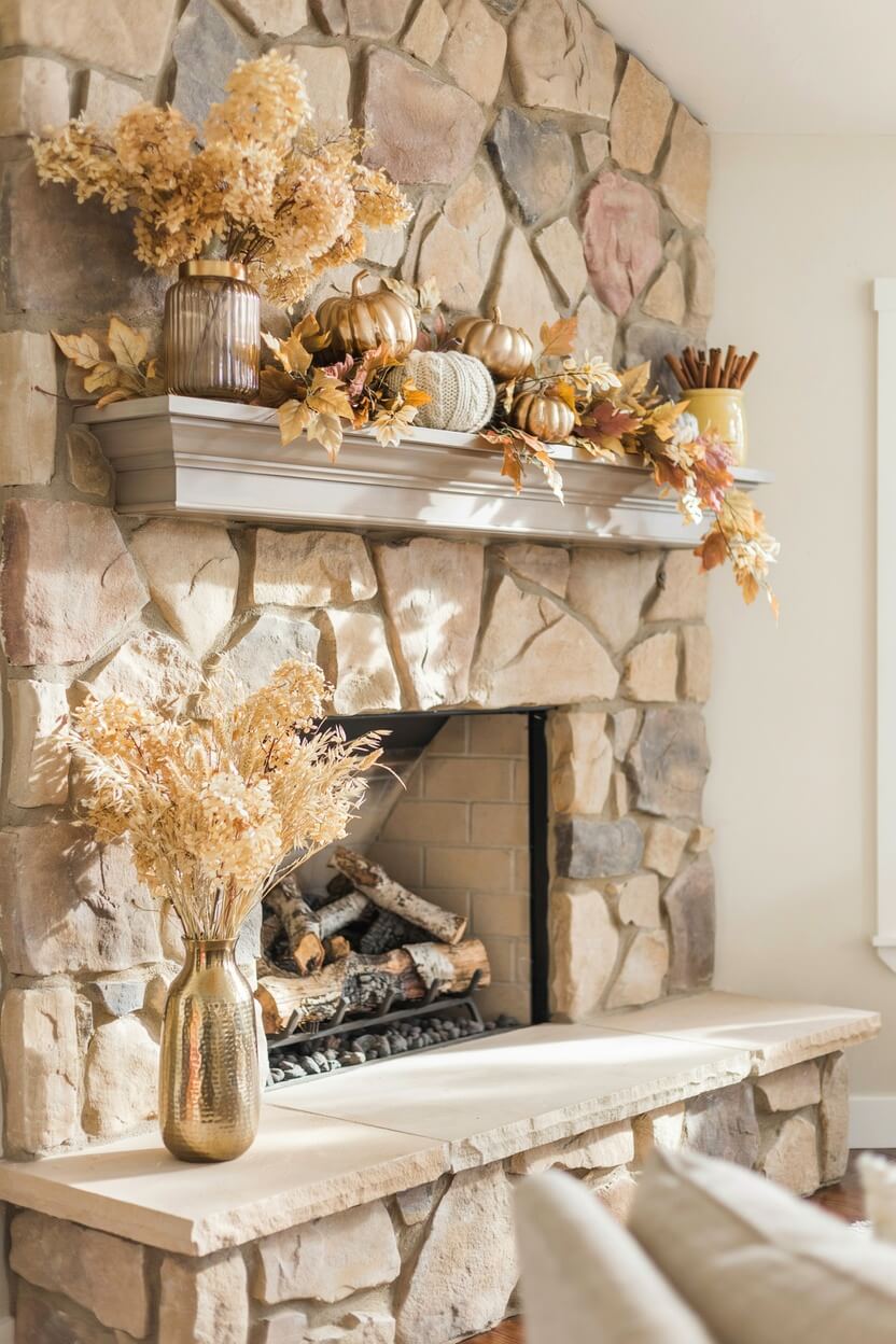 A large natural stone fireplace featuring metallic autumn mantel ideas with gold painted pumpkins, brown dried leaves, and tall tan grass in a shiny brass vase.