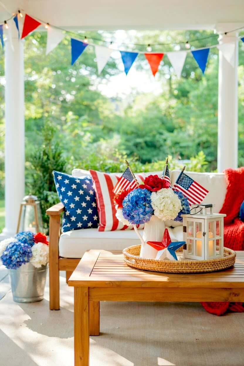 Woven tray on a wooden coffee table holding a white pitcher filled with red and blue blooms, miniature flags, and a lantern in a festive summer backyard setting.