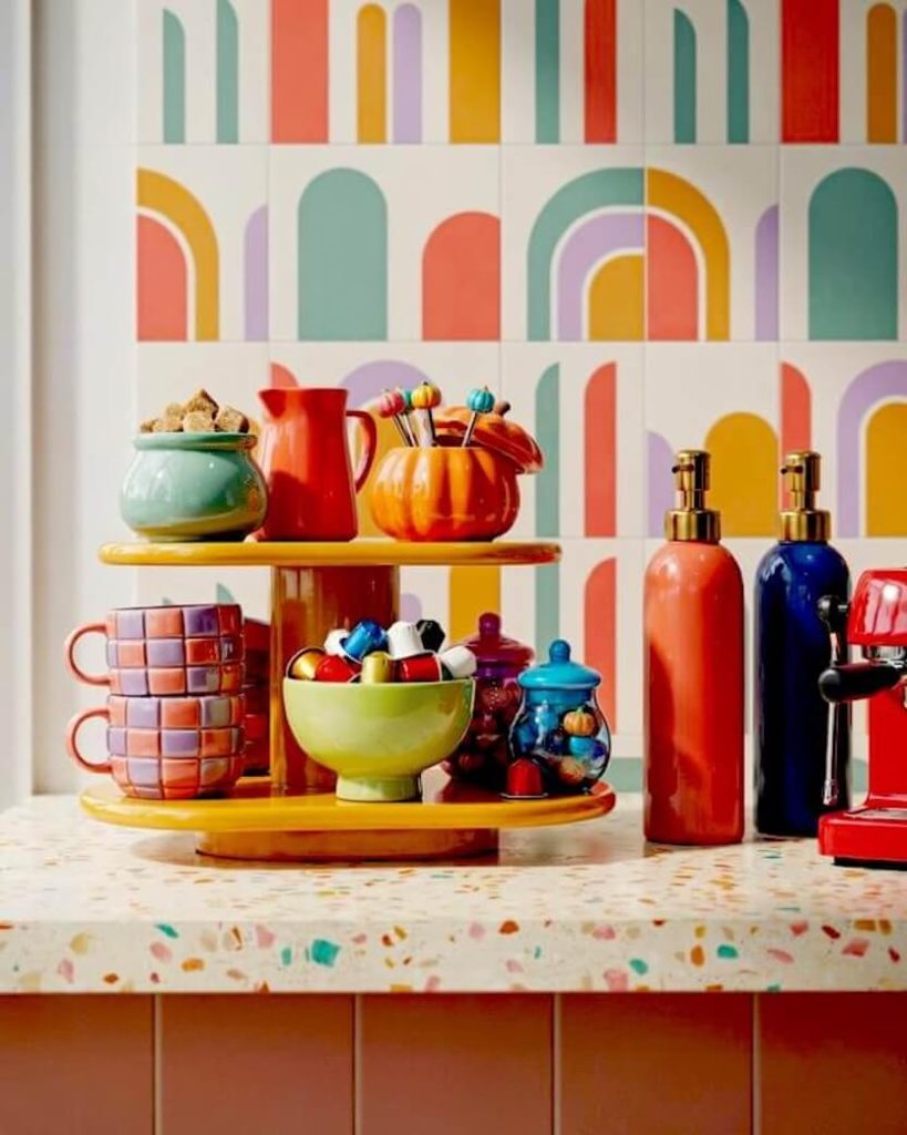 A bright yellow tray holding colorful checkered mugs, an orange pumpkin sugar bowl, and sleek coffee syrup pumps creating a retro autumn coffee bar setup.