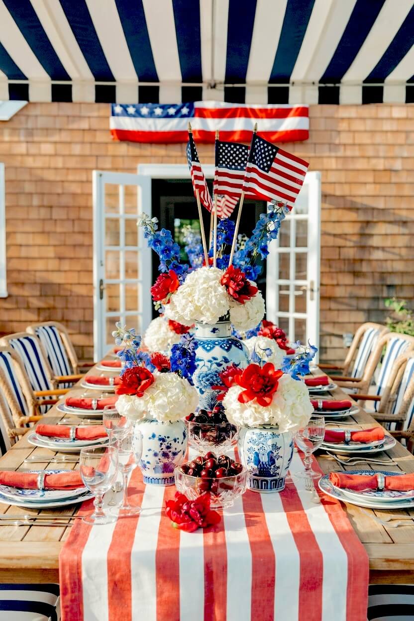 Extravagant outdoor dining setup featuring patterned ginger jars overflowing with white blooms, red flowers, and tall wooden flagsticks on a striped table runner.