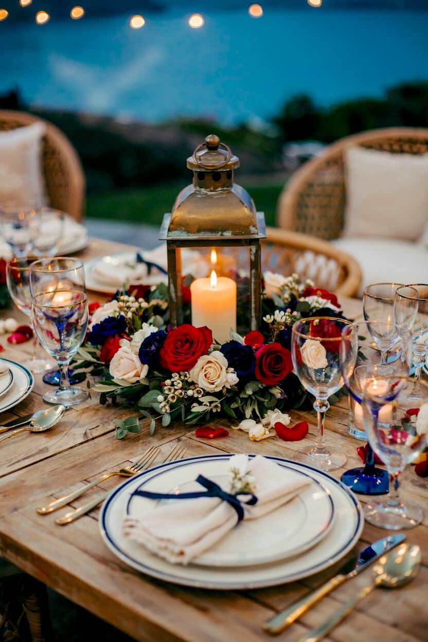 Elegant patio dining table featuring a glowing brass candle lantern surrounded by a lush wreath of dark blue roses, red blooms, and fresh greenery at twilight.
