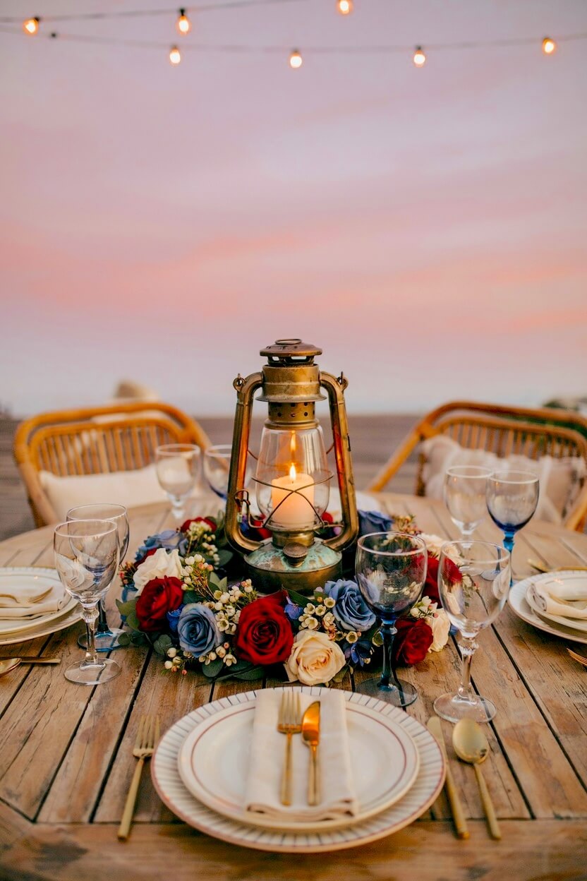 Rustic metal lantern with a lit candle resting inside a circular wreath of vibrant red, white, and blue roses on a wooden dining table against a pink sunset sky.