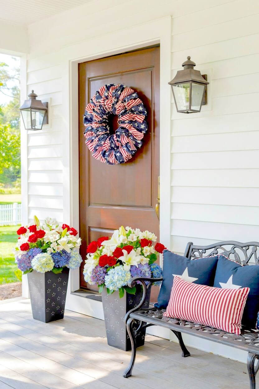 Classic front door decorated with a ruffled flag wreath flanked by tall planters filled with blue hydrangeas and red roses for summer patriotic yard styling.
