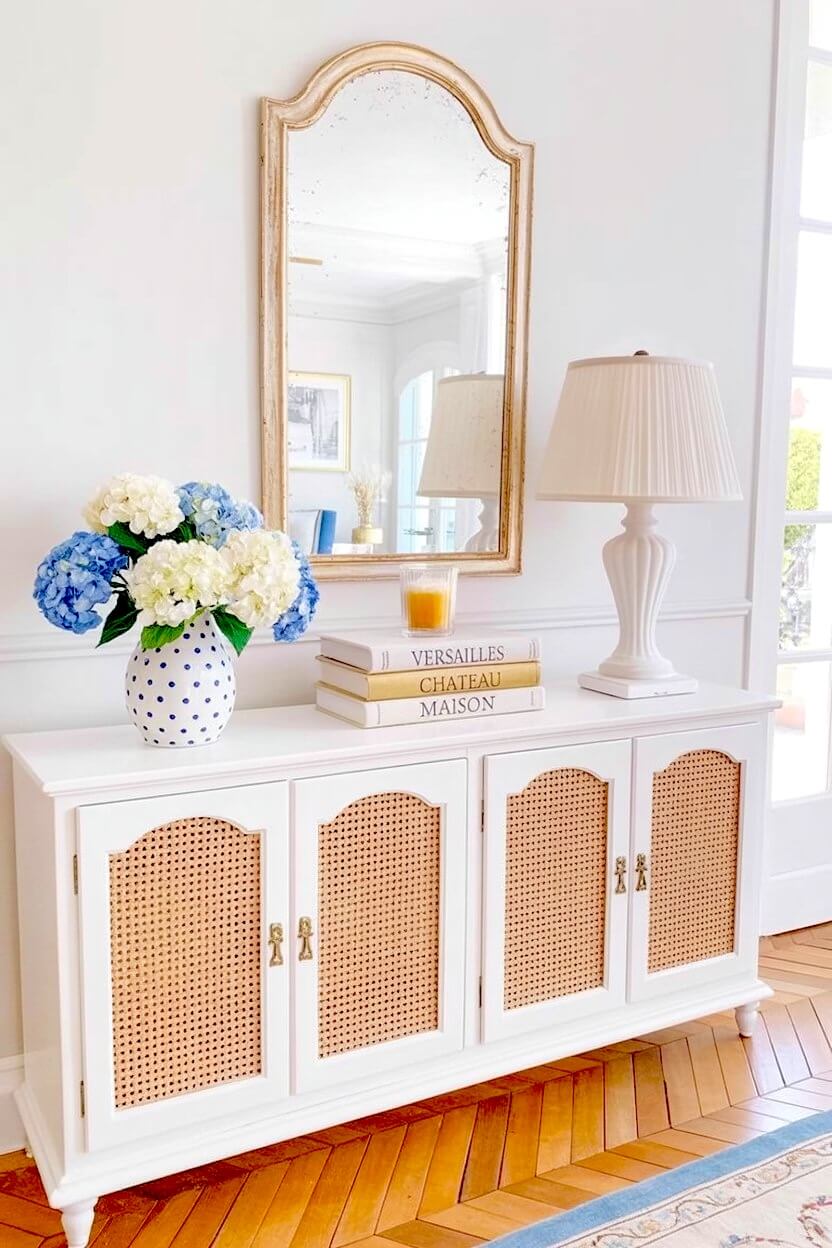 White cane console cabinet displaying vibrant blue and white hydrangeas in a polka dot vase, and a classic fluted white table lamp in a bright hallway.