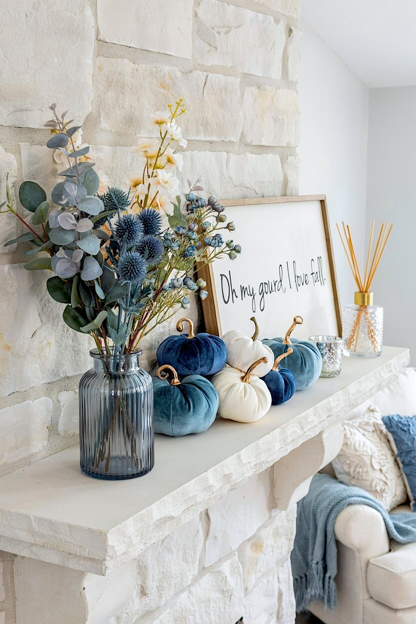 A rustic white stone fireplace highlighting coastal fall styling with a blue glass vase holding thistles, next to dark blue velvet and smooth white pumpkins.