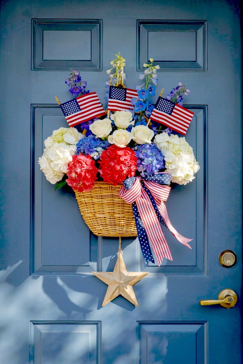 Blue painted front door featuring a woven floral basket overflowing with vibrant red and white blooms, small flags, and a wooden star for rustic patriotic styling.