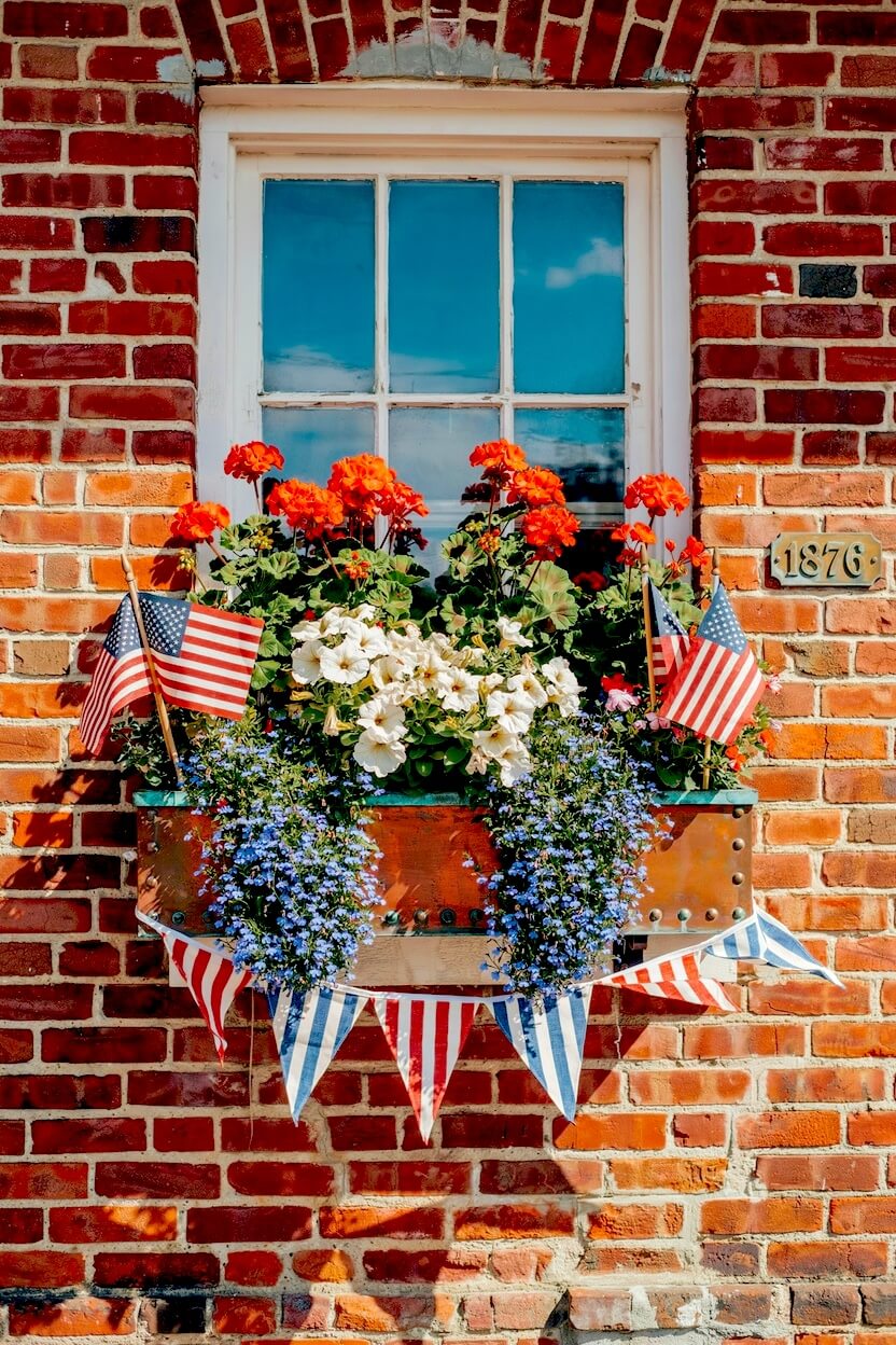 Copper window box overflowing with vibrant orange flowers, white blooms, trailing greenery, and miniature flags above a striped fabric bunting on a brick facade.