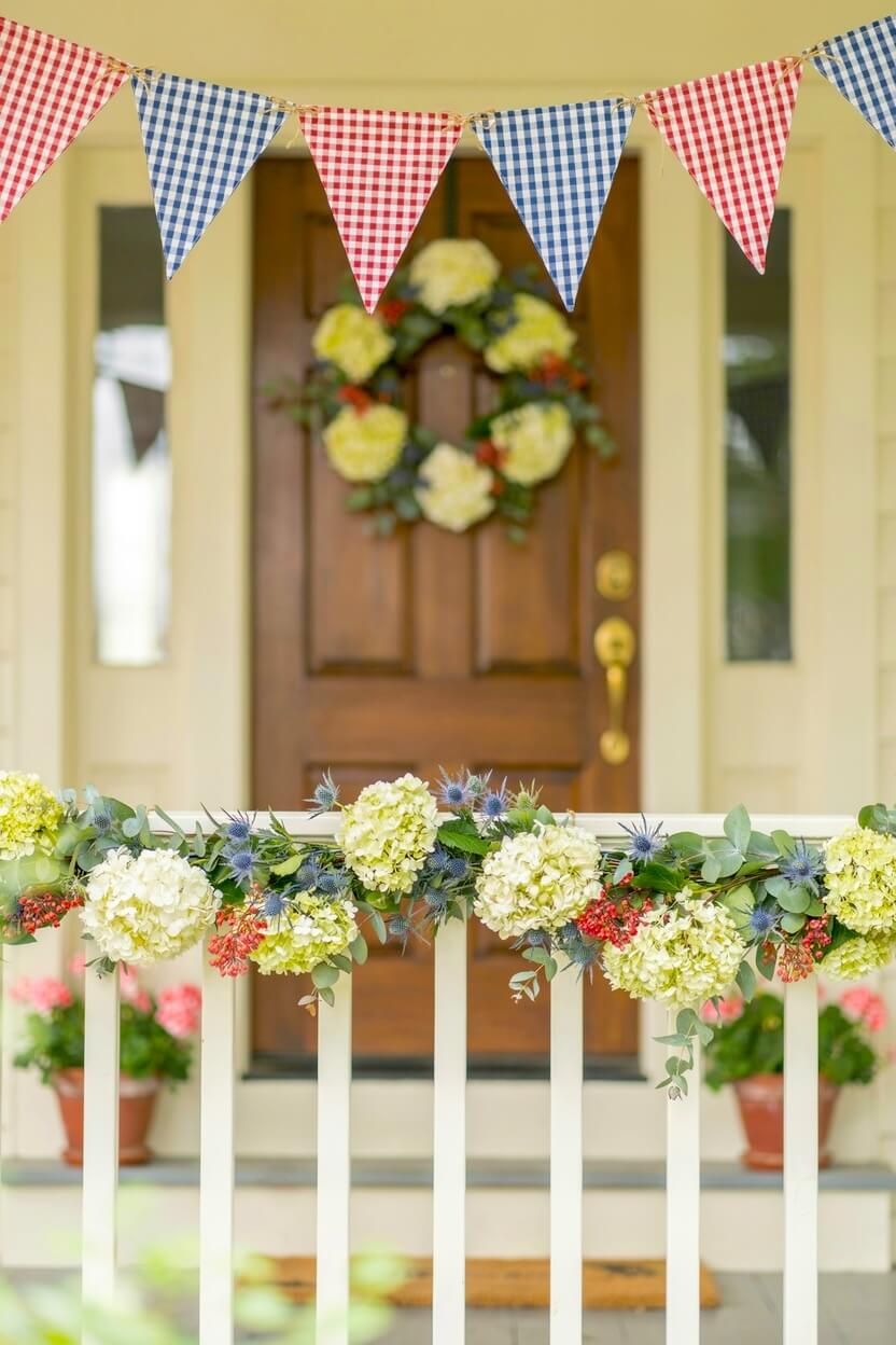 White wooden porch railing draped with a fresh green garland featuring white hydrangeas beneath a hanging red and blue gingham pennant banner for summer styling.