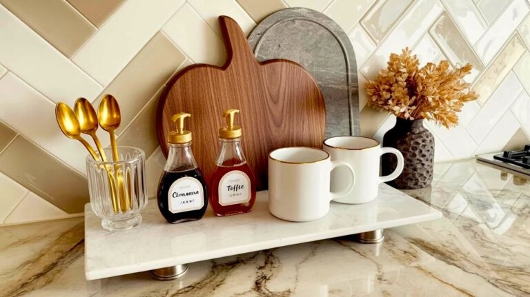 A white marble tray holds syrup dispensers, gold spoons, a wooden pumpkin board, and white mugs, presenting beautiful fall coffee corner decor ideas.