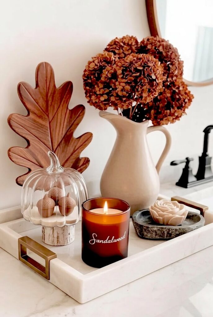 A white marble tray displays a brown glass sandalwood candle, a beige ceramic pitcher with dried brown hydrangeas, and an oak leaf for autumn bath decor.