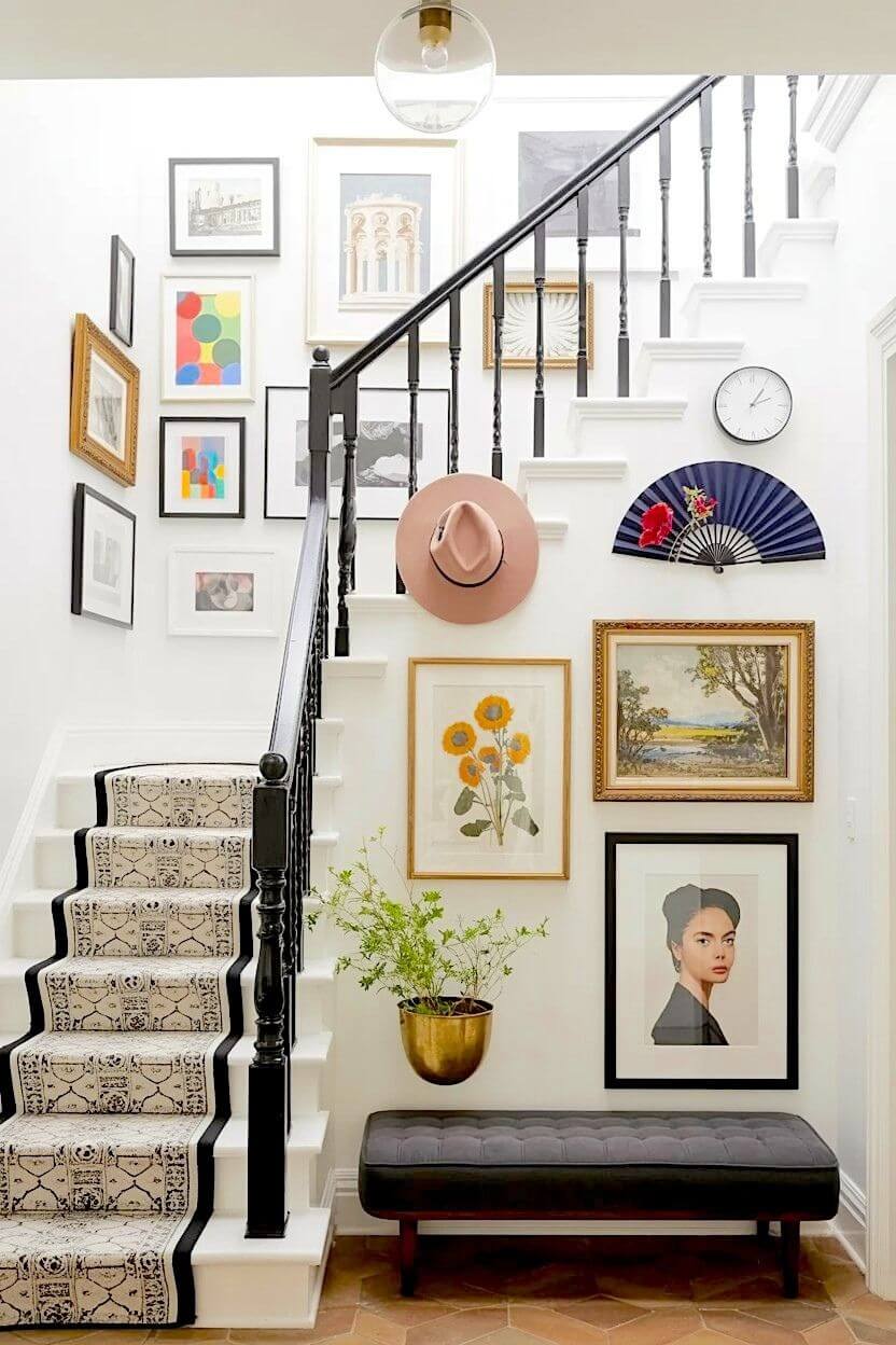 n eclectic gallery featuring a pink felt hat, a folded blue fan, vintage portraits, and landscape paintings curated as vibrant stairway wall art ideas on a white wall.