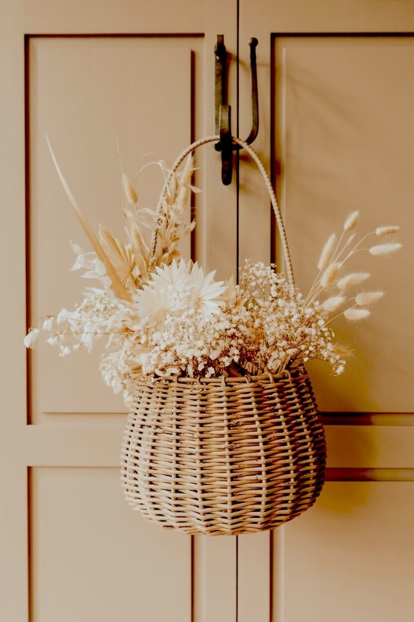 A woven wicker basket hangs from a beige cabinet door displaying a collection of dried white flowers and pale bunny tail grass for neutral autumn decor ideas.