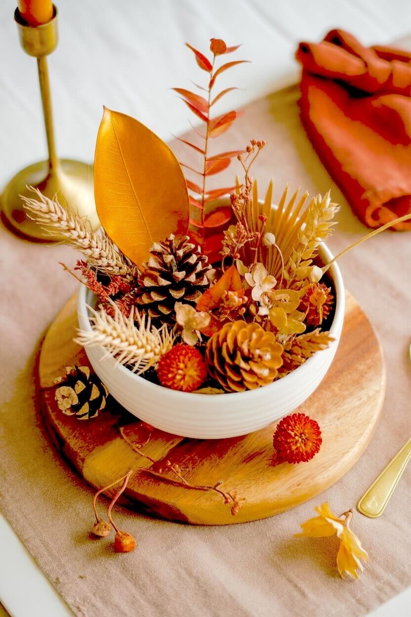 A white ceramic bowl sits on a round wooden block filled with pinecones and dried wheat stalks next to a gold leaf and orange florals for rustic fall styling.
