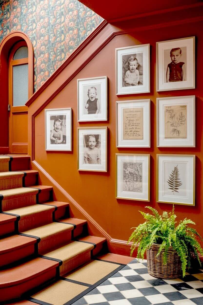 A symmetrical grid of vintage sepia children's portraits and botanical sketches in white and gold frames on a deep rust orange-painted stair wall offering design ideas.