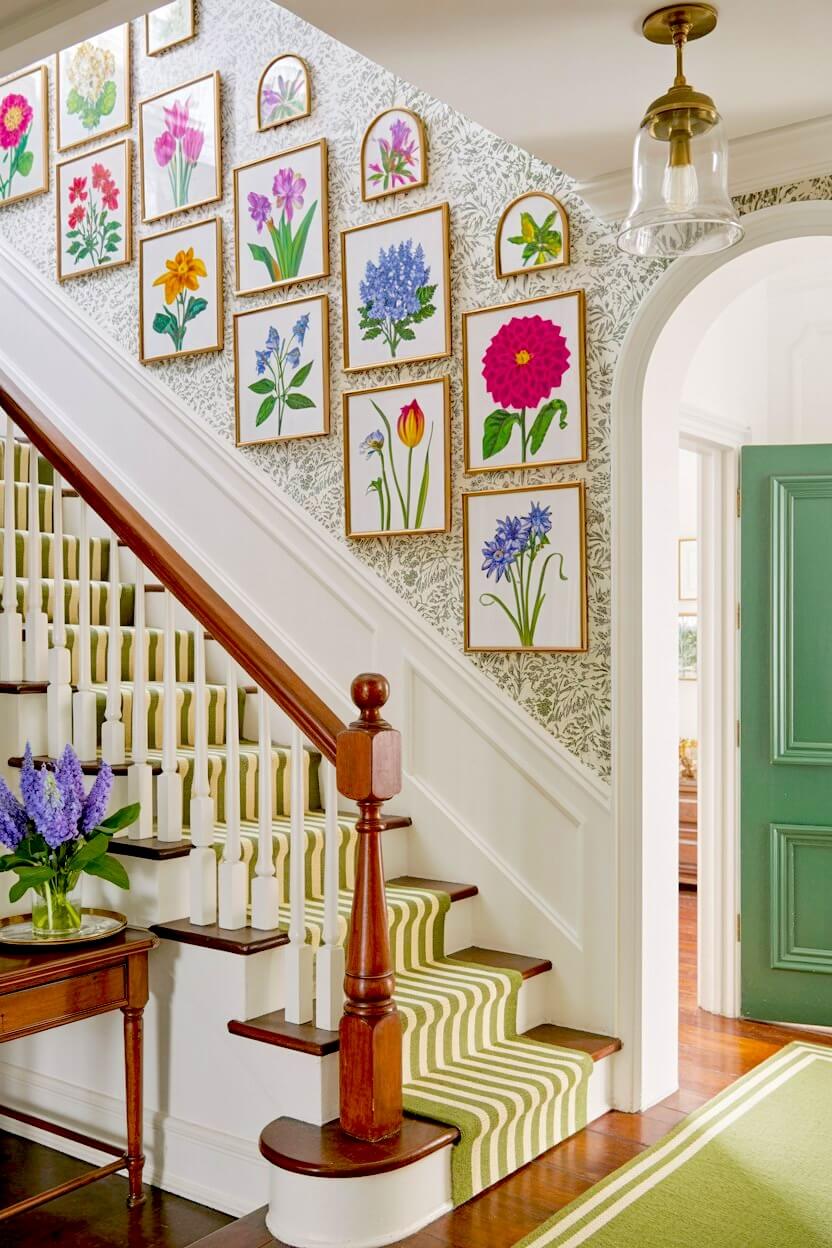 A neat grid of brightly colored floral and botanical prints in thin gold frames hanging over delicate green patterned wallpaper as traditional staircase decor.
