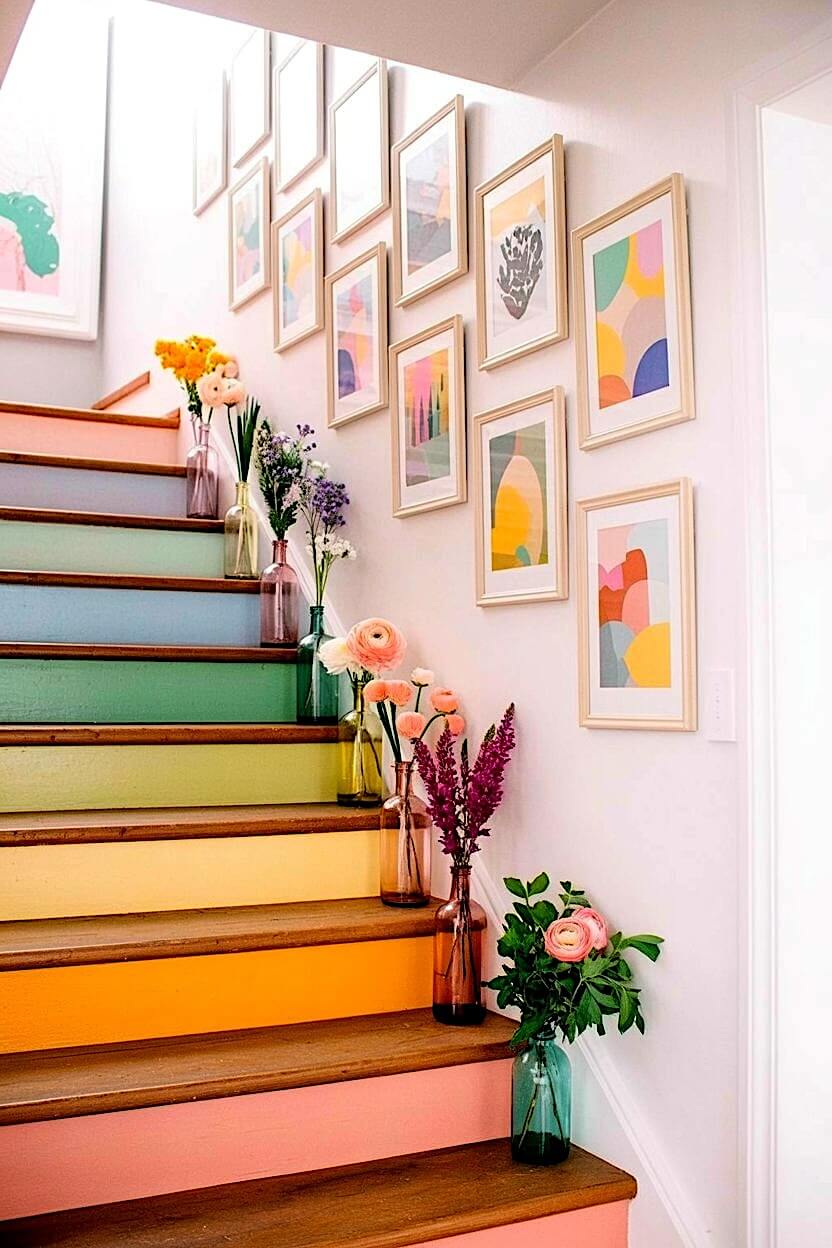 A series of abstract colorful art prints in matching light wood frames ascending a white wall beside brightly painted rainbow-colored wooden stairs with flowers.
