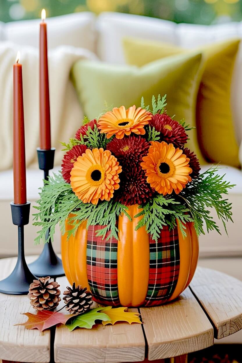 Orange plaid-wrapped traditional pumpkin flower arrangement featuring orange gerbera daisies, dark red mums, and green cedar branches on a round wooden table.